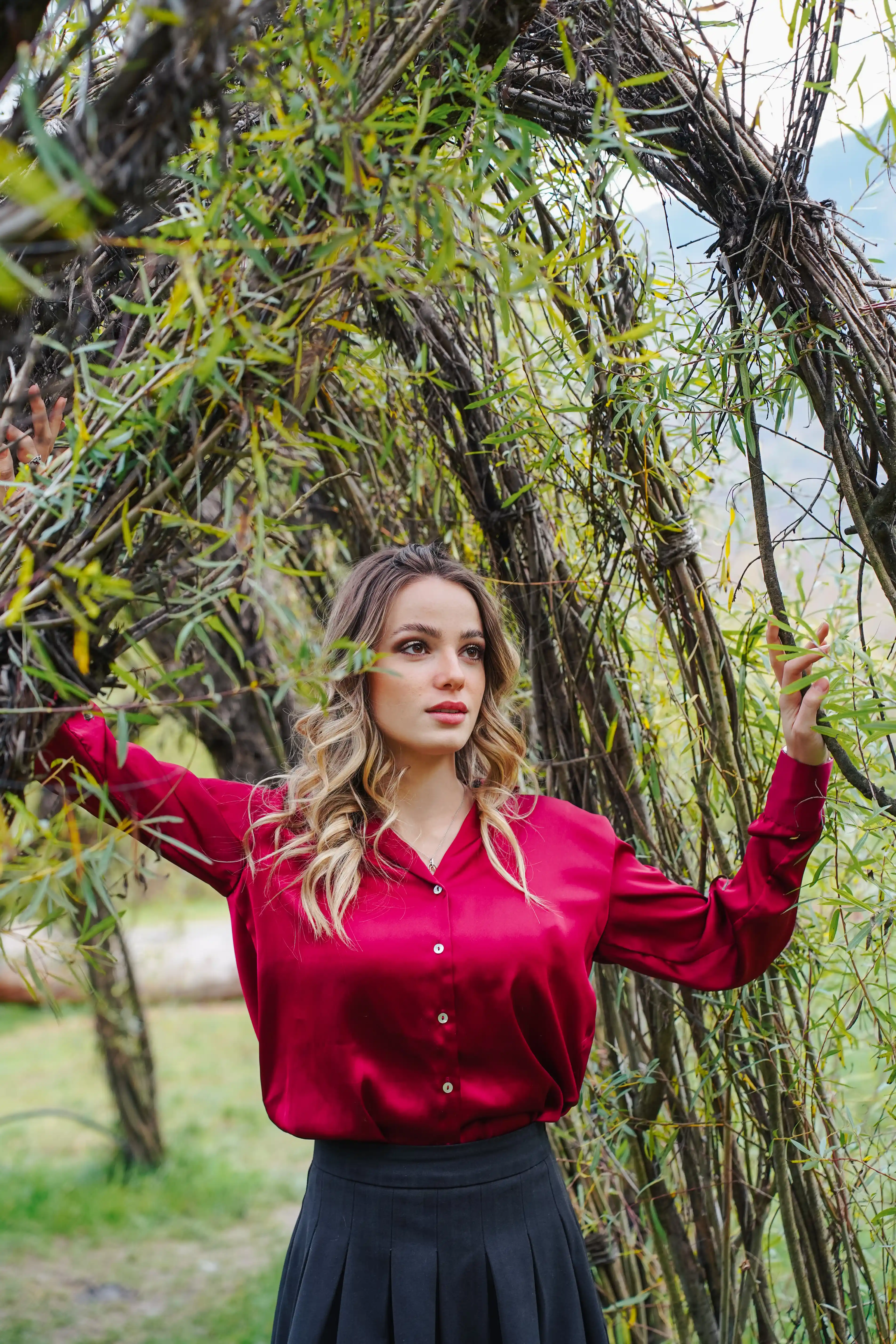 Una donna bionda con una camicia di seta rossa posa tra i rami di un salice