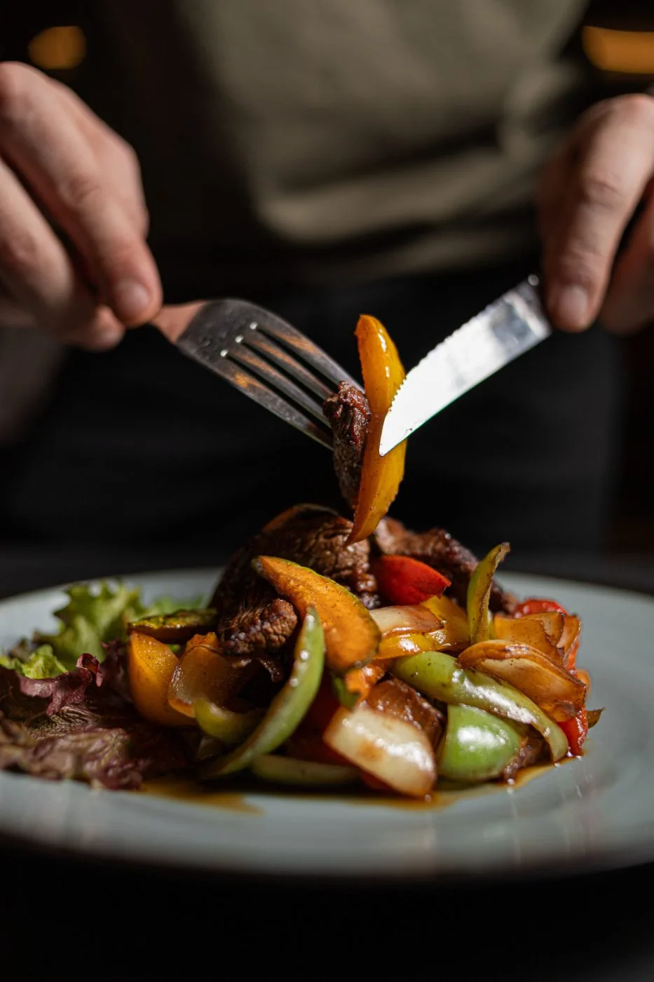 Mani che tagliano con forchetta e coltello un piatto di carne e verdure