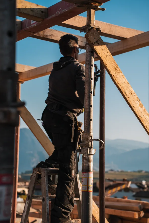 Carpentiere al lavoro su un'impalcatura in legno, con le montagne sullo sfondo.